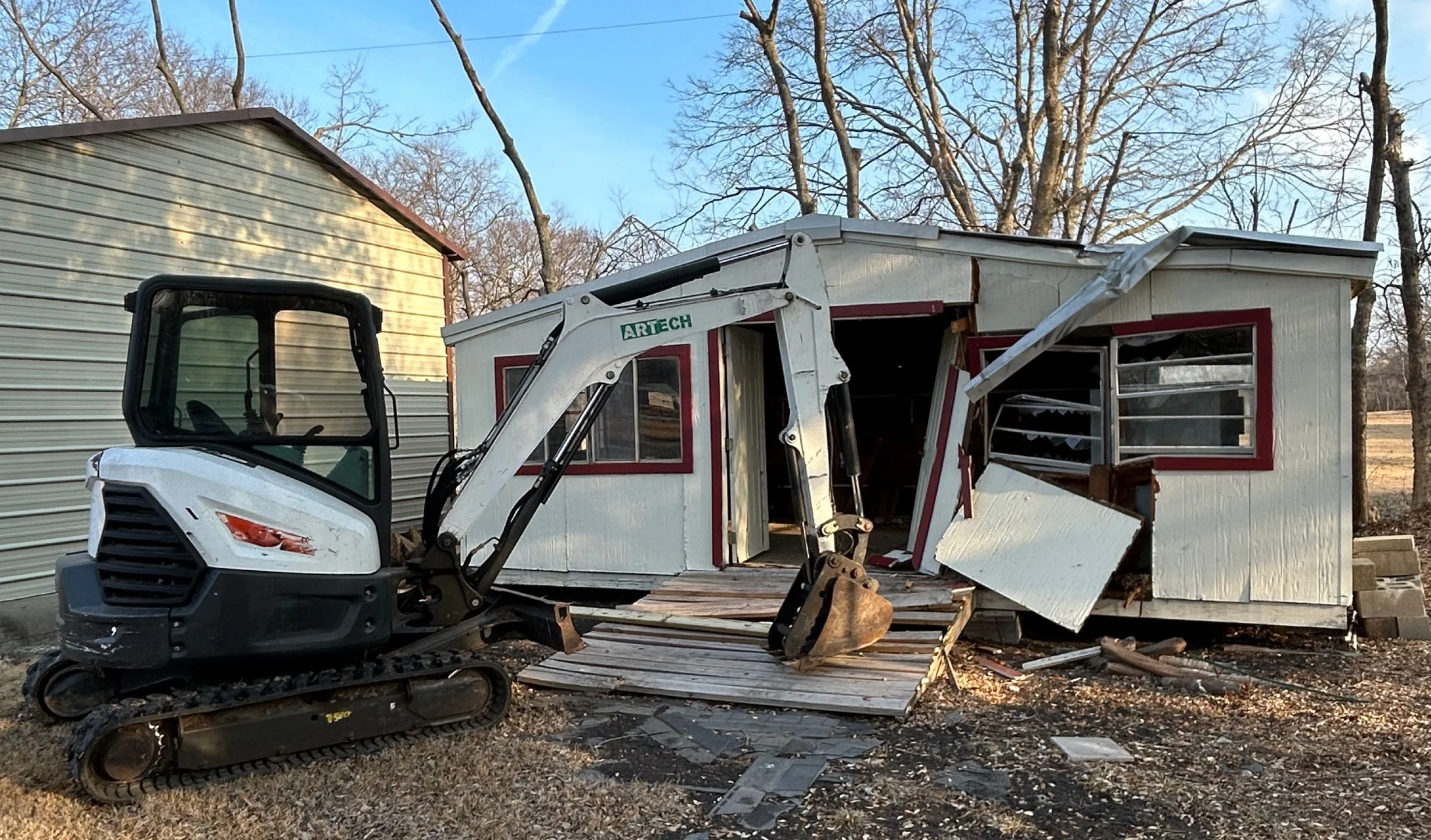 A white excavator sits next to a partially demolished building with red-framed windows under a blue sky.